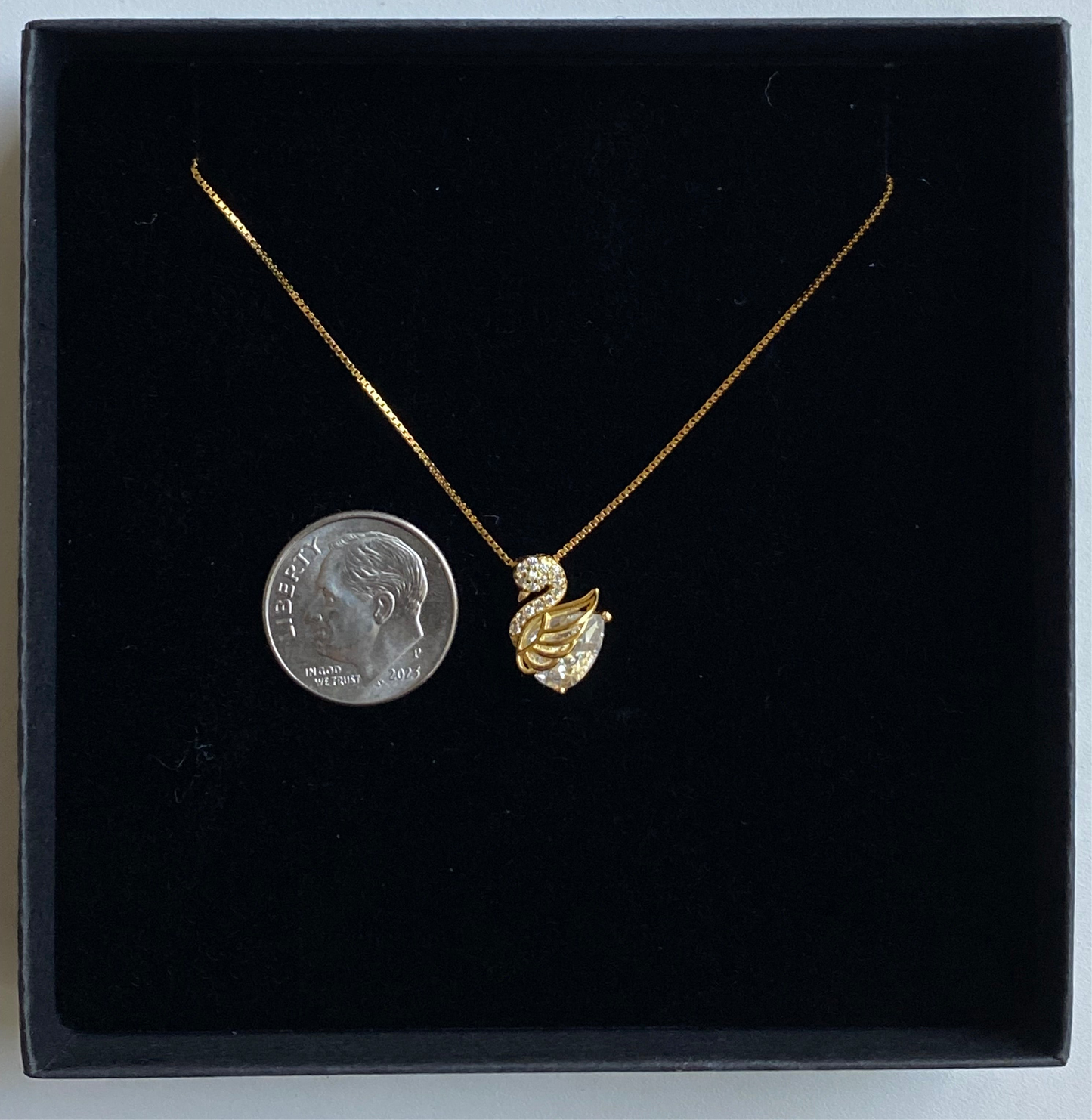 Gold necklace with a swan shaped pendant on a black background, next to a dime for scale.
