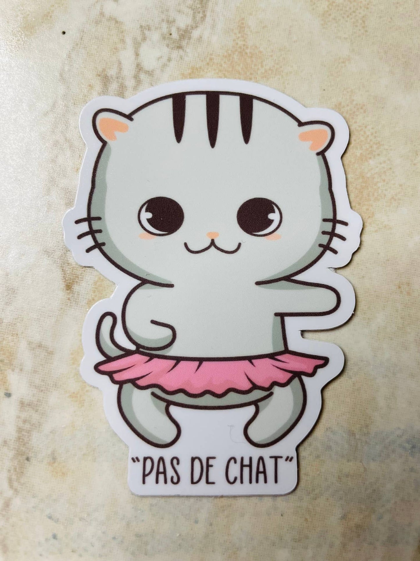 Sticker of a cat in a pink tutu with 'Pas de Chat' text on a textured beige background
