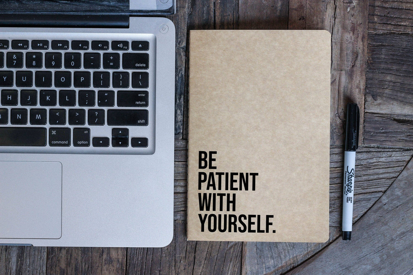Notebook with 'Be patient with yourself' on a wooden surface next to a laptop and pen.