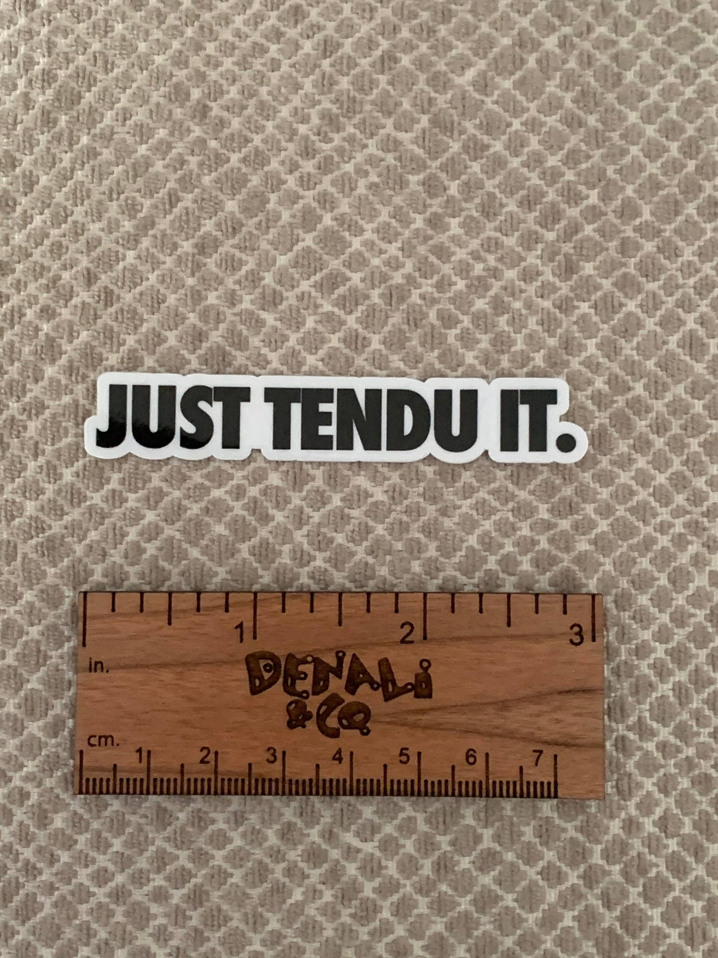 Sticker with 'JUST TENDU IT.' text on a textured beige background with a wooden ruler for scale.
