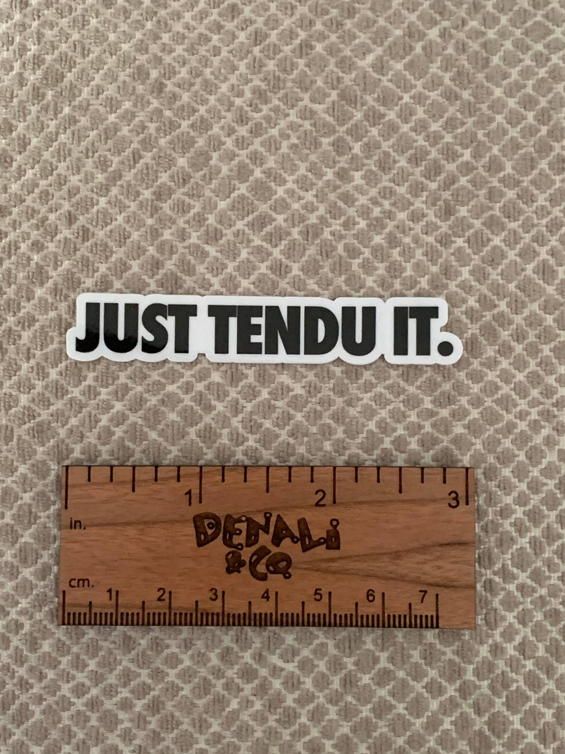 Sticker with 'JUST TENDU IT.' text on a textured beige background with a wooden ruler for scale.