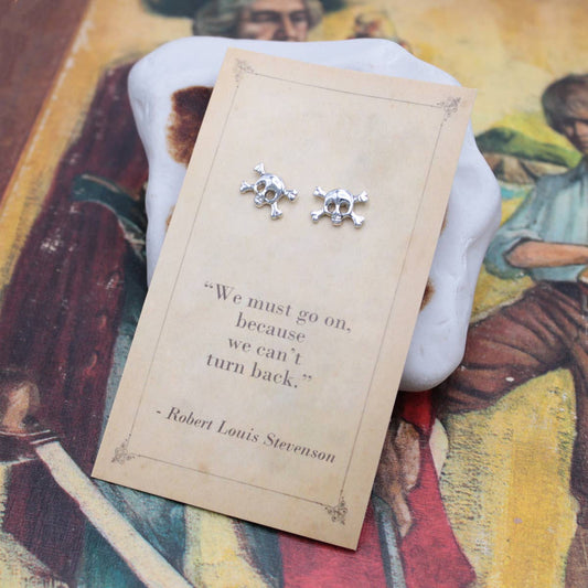 Silver skull and crossbones earrings on a card with a quote, placed on a colorful fabric background.