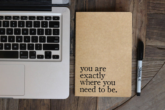 Laptop with a notebook and pen on a wooden surface, featuring an inspirational quote.