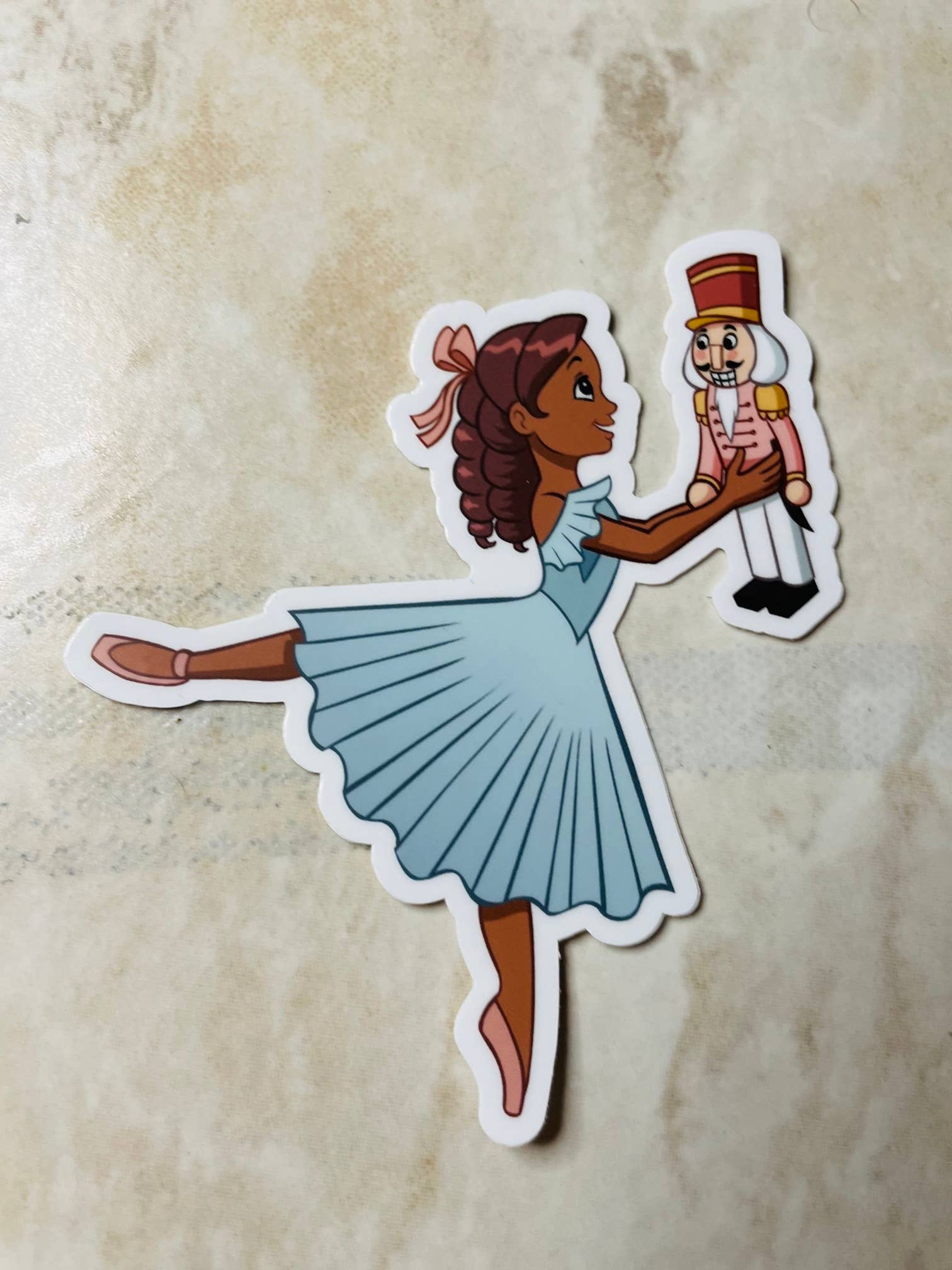 Sticker of a girl in a blue dress holding a Nutcracker toy on a textured beige background