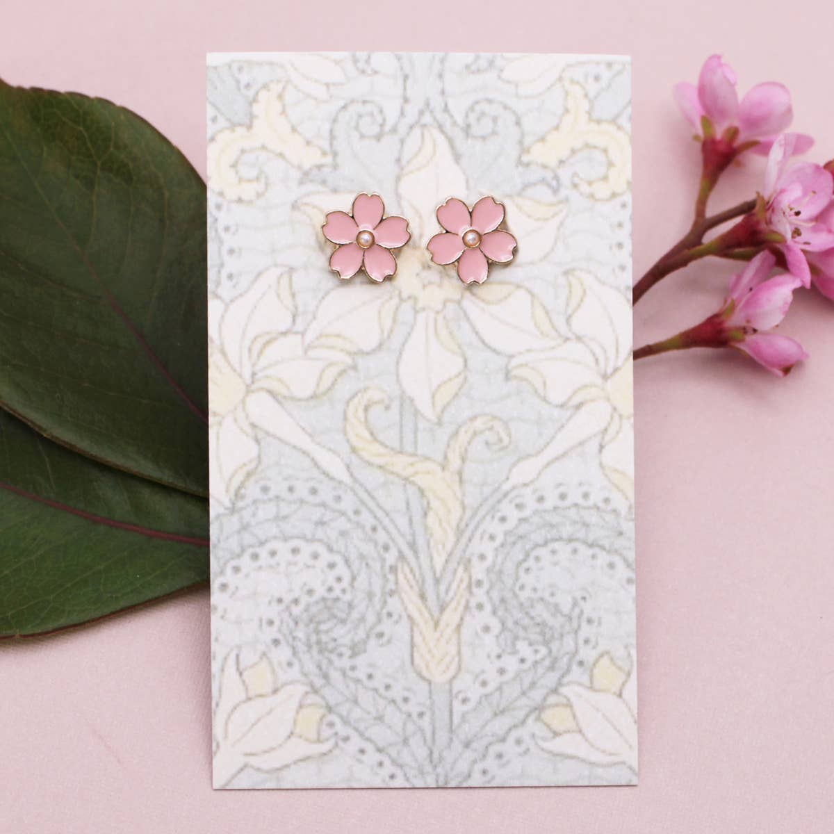 Pink floral earrings on a decorative card with floral patterns, surrounded by pink flowers and green leaves on a pink background.