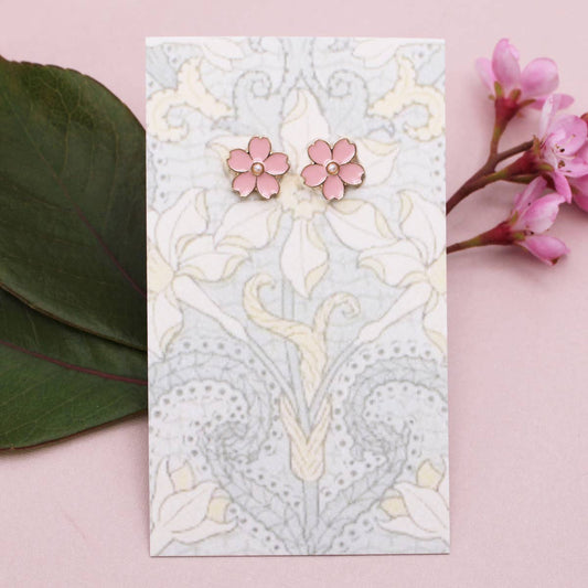 Pink floral earrings on a decorative card with floral patterns, surrounded by pink flowers and green leaves on a pink background.