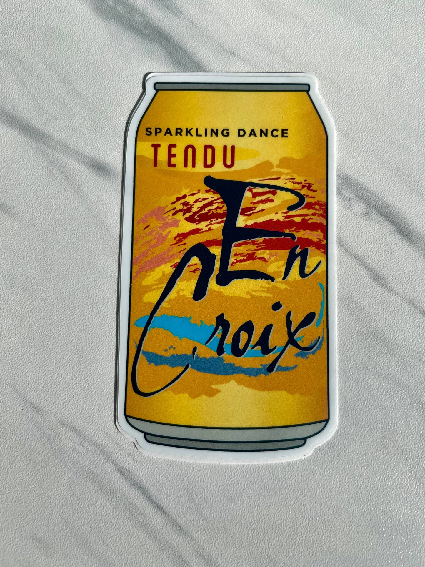 Sticker of a can with 'Tendu En Croix' branding on a gray fabric background
