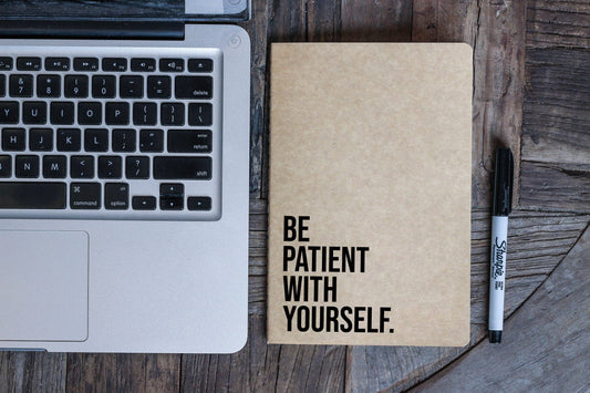 Notebook with 'Be patient with yourself' on a wooden surface next to a laptop and pen.